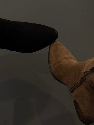 Two footwear items are centrally depicted on a dark gray, subtly reflective surface. The object in the upper-left quadrant is the pointed toe of a black shoe or boot with a smooth, matte finish. The object in the lower-right quadrant is the pointed toe and upper vamp of a brown or tan boot, appearing to be constructed from suede or a similar textured material, featuring a visible zipper running vertically along its side. The pointed tips of both footwear items are positioned in direct contact or extremely close proximity near the center of the frame. No individuals are visible. The ambient lighting is subdued. The location is Jersey City, United States.