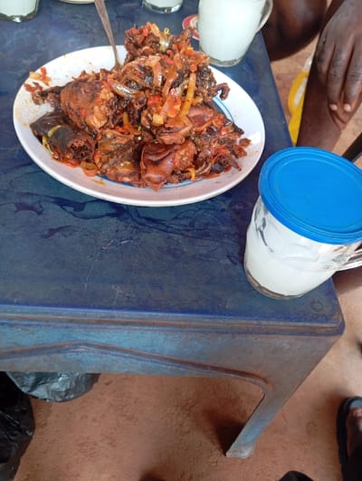 The image displays a casual meal setting, likely outdoors or in an open-air establishment within Okpatu-Ukehe, Nigeria. The specific location within the city is not visually discernible. A large white oval plate, approximately 35 cm in length, rests on a worn, dark blue plastic or metal table. The plate contains a substantial portion of a dark meat dish, possibly goat, featuring various cuts of meat with skin and bones, mixed with sliced red peppers and onions, all generously coated in a rich, reddish-brown sauce. A silver-colored metal spoon is embedded in the food.

To the right, a clear glass mug containing a milky white liquid is partially covered by a blue plastic lid. Two additional clear glasses with similar contents are partially visible in the blurred background. A dark-skinned person's right hand and forearm are visible in the upper right quadrant of the image, resting near a background glass. In the lower right, a dark-skinned foot, wearing a black sandal, is visible on the ground. The ground beneath the table is composed of reddish-brown earth, and a crumpled black plastic bag is visible on the ground to the left.