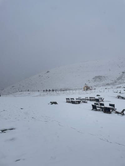 The image depicts a snow-covered mountainous landscape in Juta, Georgia, under an overcast sky with active snowfall. The foreground is entirely covered in white snow, displaying faint footprints and tracks. Multiple sets of dark wooden picnic tables and benches, partially covered in snow, are arranged in clusters across the mid-right foreground. A large, dark, snow-dusted rock or stump is present in the mid-left foreground.

In the mid-ground, a path or track runs horizontally, bordered by a low fence composed of dark posts and a thin line. Along this path, five individuals are seen riding horses from left to right. Further to the right along this same line, a multi-colored playground structure, primarily yellow and red, stands partially obscured by snow. The background consists of a snow-covered mountain slope rising steeply, with its upper reaches and the sky indistinguishable due to dense fog or low clouds, creating a hazy, grey horizon.