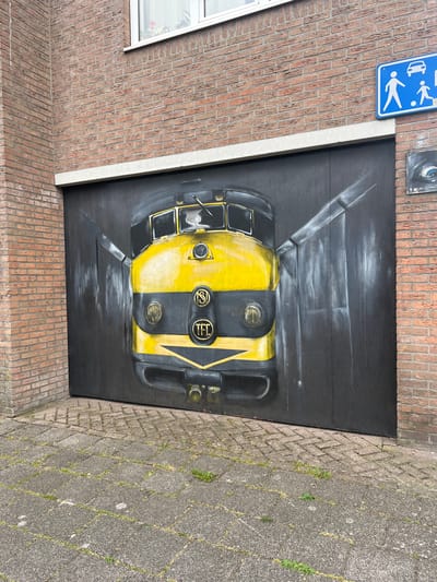 This media file features a mural painted on a black, wooden surface, possibly a door or wall, attached to a brick building. The mural depicts the front of a yellow and black vehicle, resembling a tram or train carriage, seen from a low angle looking upwards. The artwork uses a realistic style with strong contrasts between light and shadow, giving it a three-dimensional effect.
The vehicle's front is divided vertically into yellow and black sections. Prominent features include two circular headlights with visible bulbs, a windshield with wipers, and circular emblems on the black section. One emblem displays the letters "KSI" within a circular frame, and another shows "TFD" in a similar design. The mural has a dark background with streaks of white and grey, suggesting motion or light effects.
To the left of the mural, a blue sign with white pictograms is mounted on the brick wall. The sign shows a car and two people walking, indicating a pedestrian area or a warning about vehicles. A small portion of a window with a white frame and some red flowers visible inside is also on the left. The building's facade is constructed of reddish-brown brick. To the right of the mural, the ground is paved with grey interlocking bricks, with some weeds growing in the gaps. The overall scene appears to be outdoors, possibly in a public space in Leeuwarden, Netherlands, given the context. The lighting suggests daytime, but no specific time or weather conditions are discernible.