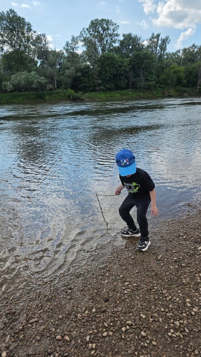 A young boy wearing a blue baseball cap and a black t-shirt with a graphic print is standing on a pebble-strewn riverbank. He is holding a stick and appears to be poking it into the shallow water. The river is calm with ripples on its surface. The background features a dense line of green trees and a cloudy sky. The scene is set in an outdoor, natural environment. The location is identified as the city of Дарьино, Russia.