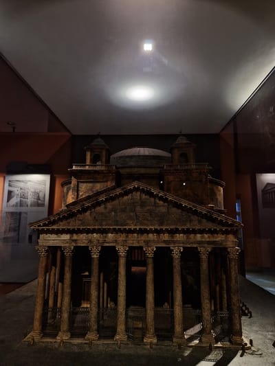 An intricate, dark bronze-toned architectural model of a classical Roman building is centrally displayed in a dimly lit indoor exhibition space in Aschaffenburg, Germany. The model features a prominent multi-columned portico with Corinthian capitals and a triangular pediment bearing a faint inscription, "M.AGRIPPA.L.F.COS.TERTIVM.FECIT." A dark metal railing or fence stands in front of the portico's columns. Behind the portico rises a large, domed rotunda, flanked on the roof by two smaller, square, multi-tiered bell-tower-like structures, each topped with a cross. The model rests on a dark, flat base. Overhead, a circular spotlight illuminates the model, casting reflections on what appears to be a glass ceiling or protective enclosure, where a grid of square light sources is also reflected. To the left of the model, a partially visible informational panel displays black-and-white architectural photographs and text against a darker wall. The environment is static, with no individuals present or active interactions depicted.