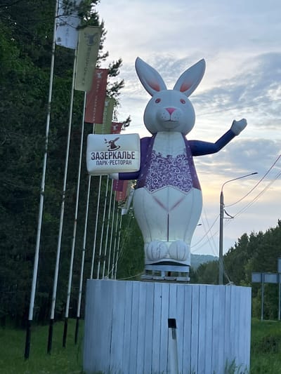 A large, inflatable sculpture of a white rabbit is displayed in front of a wooded area with a cloudy sky. The rabbit is wearing a purple vest with a white ornate pattern and holding a rectangular sign that reads "ЗАЗЕРКАЛЬЕ ПАРК-РЕСТОРАН". The sculpture is positioned on a white, wooden platform. Several flags with what appear to be symbols and text are displayed on poles to the left of the sculpture. In the background, there are trees, utility poles with wires, and a glimpse of hills or mountains. The overall impression is that of an advertisement or landmark for a park and restaurant called "Zazerkalye" in Tal'tsy, Russia.