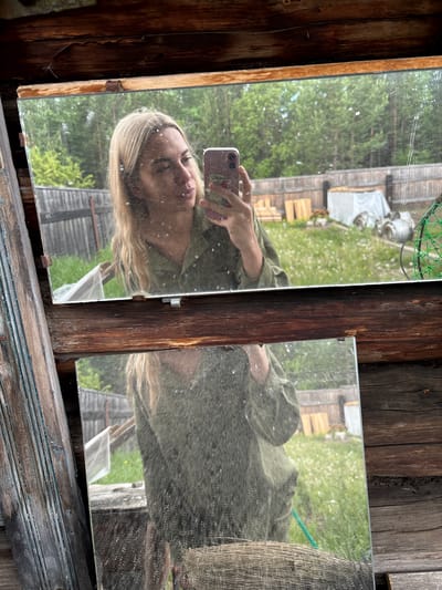 The image depicts a young woman with blonde hair taking a selfie in a mirror. She is wearing a green shirt and is holding a pink smartphone. The mirror is positioned in front of a wooden structure, possibly a shed or a rustic building. The reflection in the mirror shows a backyard with a wooden fence, green grass, and scattered white wildflowers. There are some wooden planks and a white-covered object in the background, suggesting a rural or suburban setting. The overall impression is of a casual, outdoor photograph taken in a natural environment. The location is identified as Zheleznodorozhnyi, Russia.
