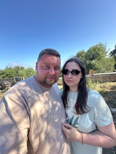 This is a selfie taken outdoors on a sunny day, likely in the afternoon, given the clear blue sky and the presence of shadows. The photo features two people, a man and a woman, in close proximity. The man is in the foreground, his face and upper torso visible, looking directly at the camera with a neutral expression. He has short hair and a beard. The woman is positioned slightly behind and to his left, also looking towards the camera. She is wearing sunglasses and has long, dark hair. She is wearing a light-colored t-shirt with a floral design. A red and gold beaded bracelet is visible on her left wrist.

The background suggests an outdoor setting, possibly a garden or patio area. There are trees and foliage visible, as well as a low wall or structure. The ground appears to be dry and covered with natural materials like dirt and small stones. The overall mood appears relaxed and casual. There is no visible text in the image. The location context is Santa Cristina d'Aro, Spain.