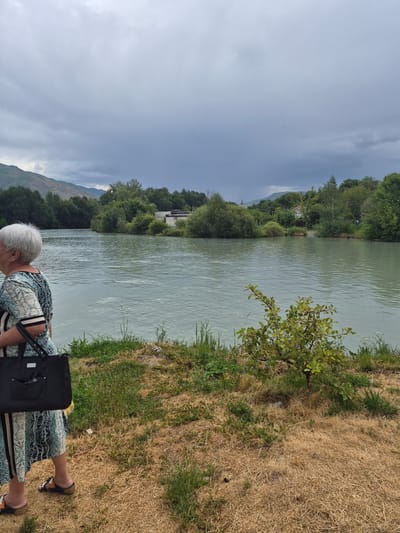 An elderly woman with gray hair stands near a riverbank, facing left and looking out at the water. She is wearing a patterned dress and carrying a black tote bag over her left shoulder. The river, with its greenish-blue water, is calm with a slight ripple on the surface. The riverbank is covered in dry grass and some green patches of vegetation. In the background, there are lush green trees and mountains under a cloudy sky. The scene appears to be in a natural setting, potentially a park or a scenic area along the river, in the city of Tekeli, Kazakhstan.