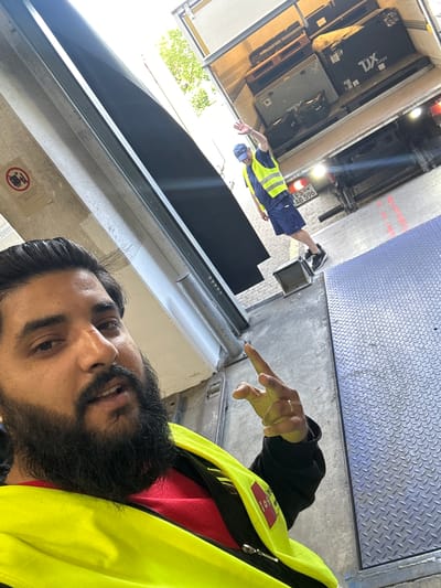 A person in a bright yellow safety vest and red shirt is posing for a selfie in a loading dock area. The person has dark hair and a beard and is making a peace sign with their right hand. Behind them, a truck is partially visible with its rear doors open, loaded with various boxes and pallets. Another person wearing a blue cap and a yellow safety vest is standing in the back of the truck, waving. The location appears to be an industrial or logistics facility, possibly in Schweitenkirchen, Germany, given the license plate on the truck which reads "FS AB 596". The ground is concrete and metal, with some red markings visible. A "no forklifts" sign is mounted on a wall to the left. The overall lighting suggests it is daytime, with sunlight filtering in.
