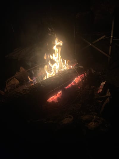 A campfire burns in a dark, outdoor environment, likely at night. The flames are bright yellow and orange, with wisps of smoke rising. The fire is contained within a ring of rocks and logs. In the background, a dark silhouette of a camping chair is visible. To the right of the fire, a red and white pack of cigarettes rests on the ground. The overall scene suggests a camping or outdoor gathering.