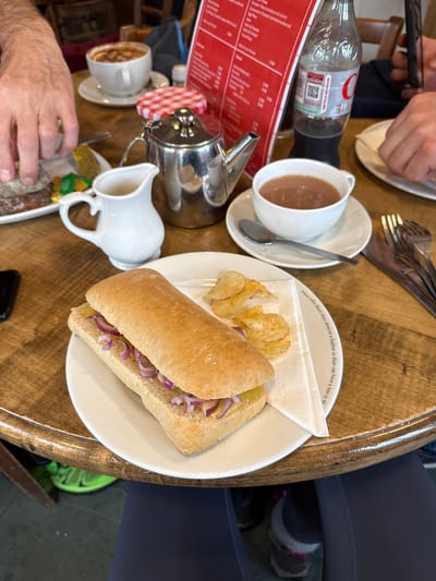 The image captures a high-angle view of a café table setting, likely in Bowness-on-Windermere, United Kingdom, based on the cafe's aesthetic. The table is made of wood. Various items are arranged, including a plate with a sandwich filled with red onions, potato chips, and a napkin, alongside a cup of tea on a saucer with a spoon. A silver teapot with a red-and-white checkered lid and a white milk jug are also present. A red menu is visible. In the background, parts of two people are visible: one with a hand reaching for the table items and another with legs wearing dark leggings and sneakers. A bottle of water and cutlery can also be seen on the table.