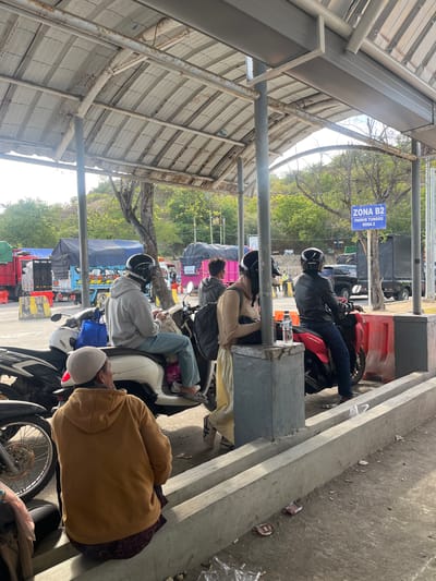 The image depicts an outdoor scene, likely a parking or waiting area, under a covered structure. Several people are present, some seated on motorcycles and others standing. In the foreground, a person wearing a brown hooded sweatshirt sits on a low concrete barrier, facing away from the camera. To their left, the wheel and front of a black motorcycle are visible. To the right, two individuals are seated on white and red motorcycles, respectively. The person on the white motorcycle wears a grey hoodie and blue jeans, while the person on the red motorcycle wears a black leather jacket and blue jeans. Both are wearing helmets. Further back, other individuals and several trucks, some with tarpaulin covers, are visible, suggesting a transportation hub or a point of transit. A blue sign with white text stating "ZONA B2 PARKIR TUNGGU RODA 2" is mounted on a pole to the right. The environment appears somewhat weathered, with exposed metal structures and concrete barriers. The ground is paved and shows signs of debris. The overall impression is one of a temporary waiting area, possibly at a ferry terminal or a border crossing, given the presence of trucks and multiple individuals on motorcycles. The context of Upt Tambak Sari, Indonesia, suggests this is a location within that administrative area, likely a public facility.