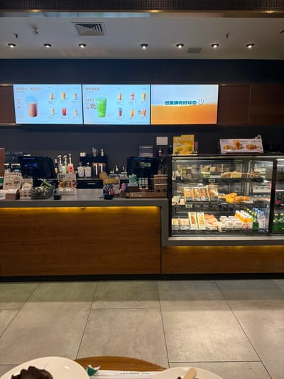 The image captures the interior of a brightly lit cafe, likely located in Yuhuatai District, China. The primary subject is a service counter with a prominent display case filled with various food and beverage items, including sandwiches, pastries, and bottled drinks like Evian water. Behind the counter, shelves hold additional products and equipment.
To the left of the counter, several vertical digital menu boards showcase a selection of drinks. One large screen displays Chinese text, "想要拥有好状态" (Want to have a good state/condition), accompanied by a smiling emoji. Below it, other screens list coffee and tea options with prices. Visible text includes "咖啡时光, 低因可选" (Coffee time, decaf optional), "Enjoy Your Coffee," "咖啡师推荐" (Barista recommendations), "Barista favorites," and specific drinks like "摩卡" (Mocha - 36), "抹茶拿铁" (Matcha Latte - 29), "高蛋白拿铁" (High Protein Latte - 36), "红茶拿铁" (Black Tea Latte - 39), "美式咖啡" (Americano - 29), and "拿铁" (Latte - 33). The Starbucks logo is visible on some promotional materials near the display case, along with items like "慢煮芝士蛋" (Slow-cooked Cheese Egg) and "高蛋白拿铁牛肉意式千层" (High Protein Latte Beef Italian Lasagna).
The scene is illuminated by overhead lights and strip lighting along the counter, creating a warm ambiance. The ceiling features recessed spotlights and an air vent. The floor and a wall to the right are covered in large, light-grey tiles. No people are visible, suggesting the image might have been taken during a quiet moment or focused on the cafe's offerings. The overall impression is that of a modern, well-stocked cafe ready to serve customers.