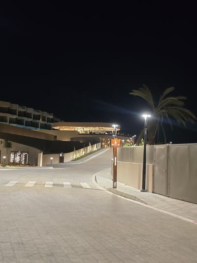The image shows an exterior view of a modern building complex at night in Polis Chrysochous, Cyprus. The architecture features multiple levels with balconies and large windows, illuminated by interior lights. A gently curving paved driveway leads towards the building, marked with a pedestrian crosswalk. A tall palm tree stands to the right of the driveway, silhouetted against the dark sky. A lamppost with a modern design illuminates the immediate area. A wooden signpost with pictograms and text, potentially indicating directions or services, is visible on the right side of the walkway. The ground is paved with interlocking grey tiles. The overall impression is one of a well-lit, upscale establishment, possibly a hotel or resort, with a clean and contemporary aesthetic.