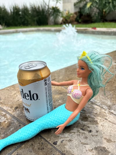 The image depicts a mermaid doll with light blue hair and a seashell bikini top, lying next to a can of Modelo Especial beer on a stone surface. The mermaid doll's tail is light blue and scaly. The can of beer has condensation on it. In the background, there is a body of water, possibly a pool, with a fountain feature. A blurred view of green foliage is visible on the left side of the image. The OCR data on the can indicates the product is brewed in Mexico by Cerveceria Modelo de México. The phrase "DE ESTE PRODUCTO ES NOCIVO PARA LA SALUD" appears in Spanish, which translates to "THIS PRODUCT IS HARMFUL TO YOUR HEALTH."