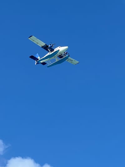 A seaplane, primarily white with blue accents and blue pontoons, is captured in mid-air against a clear blue sky. The aircraft is angled, with its tail pointing towards the upper left of the frame. A registration number, "Q-RAB", is visible on the wing. The seaplane appears to be flying over the ocean, which is not visible in the frame. The image was likely taken from below, looking up at the aircraft. This type of seaplane is commonly used for transport between islands in the Maldives, including services to and from Malé.