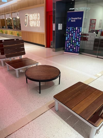The image depicts an indoor public space, likely a shopping center or mall, featuring a bank and a food establishment, with a seating area in the foreground. No people are visible, suggesting the area might be quiet or outside peak hours.

On the left side of the frame, there is a glass-fronted bank with an interior reception area visible. A prominent blue banner stands in front of the bank's entrance. The banner displays the text "PeopleFirst Bank" in white, alongside a colorful geometric pattern composed of various shapes like semi-circles, rectangles, and lines in shades of purple, blue, green, and white.

In the center and foreground, there's a seating area comprising several tables and benches. Two styles of tables are present: one round table and at least two square tables. All tables and benches feature wooden slatted tops and white or black metal frames. They are arranged on a light-colored, possibly terrazzo, floor with subtle white and light-yellow linear patterns.

Towards the bottom right, another establishment is visible, identified by an illuminated sign with the text "SHINSEN EXPRESS" in a neon-style font. The facade of this establishment features a light wooden paneling and slat design.

The lighting in the scene is bright and even, characteristic of an indoor commercial environment during daytime. The overall impression is one of a clean, modern, and unoccupied public space. The location context provided is Indooroopilly, Australia.