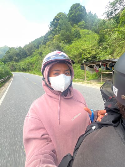 Two individuals are riding on a motorcycle on a paved road in a rural mountainous area, likely in Bình Lư, Vietnam. The person in the foreground, wearing a pink hoodie and a white face mask, is looking at the camera. They have a pink and white helmet on. Behind them, another person, wearing a black helmet, is visible from the back. The road curves to the left, bordered by a guardrail on the left and lush green vegetation and trees on the right. A small, makeshift shelter with clothes hanging on it is visible on the right side of the road. The sky is bright and clear with some clouds. The overall scene depicts a journey through a scenic, natural landscape.