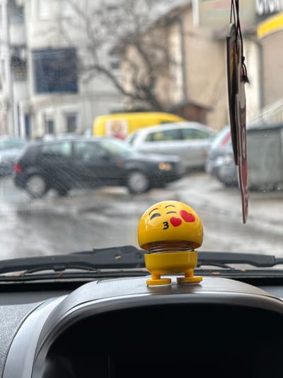 The image depicts an urban street scene in Kumanovo, North Macedonia, viewed from inside a vehicle, likely through a front windshield. In the immediate foreground, a yellow emoji-themed bobblehead with closed eyes, puckered lips, and two red hearts on its cheek is positioned on a dark gray vehicle dashboard. The bobblehead's head is mounted on a visible spring. To the right, a dark, rectangular air freshener hangs from an unseen point above. The windshield itself shows slight scratching or residue.

Through the windshield, a wet, paved street is visible, suggesting recent rain or snowmelt. Several vehicles are present: a black compact car is centrally positioned, either parked or moving slowly. Further down the street on the right, a yellow commercial van with a partially visible logo is parked. Other blurred vehicles, including a silver car, are in the background. Multi-story buildings, typical of urban architecture, line the street, with a bare tree visible amongst them. The sky is overcast. No individuals are directly visible.