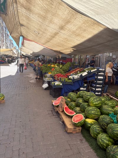 A street market in Zeytinburnu, Turkey, is depicted in the image, showcasing an outdoor setting under a large, taut canvas awning. The ground is paved with interlocking brick pavers. To the left, a building with multiple stories and numerous windows lines the street. The market stalls are laden with a variety of fresh produce, including watermelons, tomatoes, lettuce, and other vegetables. Several people are visible, some appearing to be vendors and others shoppers, engaged in their daily activities. The scene suggests a bustling, vibrant marketplace atmosphere.