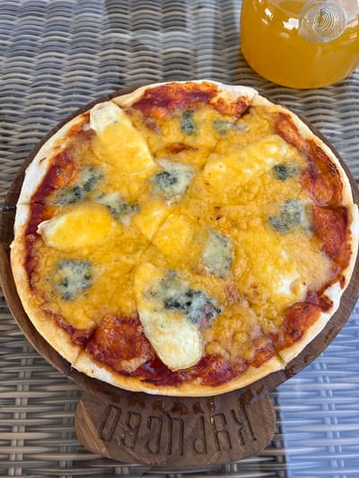 An overhead shot captures a small, round pizza on a wooden board with a carved inscription. The pizza is topped with melted cheese, including a yellow variety and pieces of blue cheese, all on a bed of red sauce. The crust is lightly browned. To the upper right of the pizza is a clear glass bottle filled with an orange beverage, possibly juice, with a silver coil-like object inside. The pizza and bottle rest on a woven, wicker-like surface, likely a table. The inscription on the wooden board appears to be "KRPUGOV". The scene is set outdoors, suggested by the diffused natural lighting. No people are visible in the image.