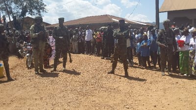The scene takes place in Kirumba, Democratic Republic of Congo, at the coordinates -0.56 latitude and 29.179 longitude. There are over 50 people gathered in an open dirt area. The people are a mix of men, women and children, some of whom are in military fatigues. There are at least three armed military personnel, one holding a rifle in his right hand, another holding a rifle in his left hand, and another holding a rifle in his right hand. The military personnel are standing in front of the crowd, while the rest of the crowd is standing behind them, facing the military personnel. Several motorcycles are parked to the left of the image, and a single-story building is visible in the background. There is a large, white sign behind the military personnel, but it is unclear what it says. The ground is a mix of dirt and small rocks. The sky is blue with a few clouds. The scene is likely a public gathering, possibly a demonstration or a meeting. The military personnel are likely in a security role.