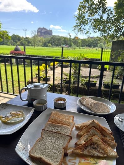 The image depicts a breakfast setting in Sigiriya, Sri Lanka. A table is set outdoors with a view of green fields and a distant rock formation, likely Sigiriya Rock. On the table are plates of toast (some triangular), thin pancakes, a small pot of jam, a teapot, a cup, and a fried egg. A metal railing separates the table from the view. The sky is blue with some clouds.