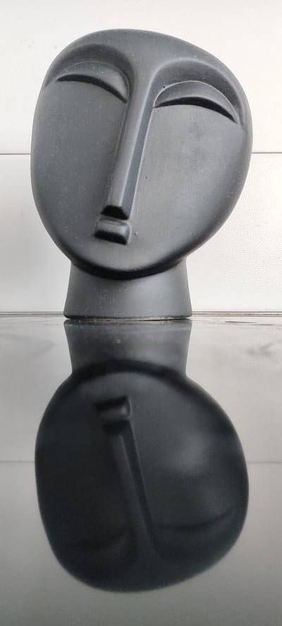 A black, abstract sculpture of a human head sits on a reflective black surface. The sculpture has stylized, almond-shaped eyes, a prominent, angular nose, and a small, rectangular mouth. The background consists of a white wall with a subtle, circular patterned texture. The sculpture's reflection is clearly visible on the polished surface below. The image was taken in an indoor setting in Snezhinsk, Russia.