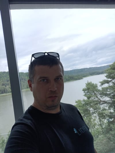 A man is taking a selfie indoors, looking at the camera. He has short, dark hair and is wearing sunglasses on top of his head. He is wearing a black t-shirt with a light blue logo on the left side of his chest. Behind him, through a large window, a scenic view of a body of water surrounded by lush green trees and hills can be seen under a cloudy sky. This appears to be the Mácha Lake region near Doksy, Czechia, given the distinctive landscape. The man is positioned in the foreground, with the natural scenery filling the background.