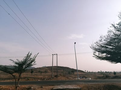 The image shows a rural scene in Rafin Pa, Nigeria. A paved road runs horizontally across the bottom of the frame, with patches of exposed brick. Beyond the road is a dry, open landscape sparsely populated with trees and shrubs. A small hill rises in the center of the frame, also sparsely vegetated. Utility poles and power lines stretch across the scene from left to right. A single streetlight stands near the center of the frame. A tree is partially visible on either side of the frame. The sky is overcast and pale.