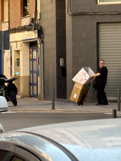 A person wearing glasses and a black outfit, including black pants and a black t-shirt, is seen from the waist up, looking towards the camera. They are standing on a paved surface next to a grey building with horizontal ribbed siding. The person is pulling a rolling cart loaded with two cardboard boxes. The larger box on top is white with red lettering that reads "WELCOME" and displays an image of a circular object, possibly a faucet or appliance. Below it is a smaller, brown cardboard box. The scene appears to be outdoors, with a building facade visible behind the person. The lighting suggests it is daytime, possibly late afternoon given the long shadows. The overall setting seems to be an urban or commercial area. The person's expression is neutral, and they are engaged in moving the boxes, implying a delivery or moving activity. The image is oriented vertically, with the ground appearing on the right side of the frame.