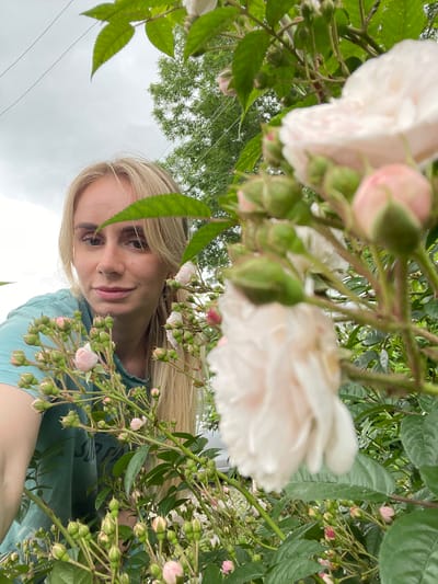A blonde woman with long hair, wearing a teal t-shirt, is positioned in the foreground of the image, partially obscured by a rose bush with light pink and white blooms and numerous green buds. She is looking towards the camera with a slight smile. The background features green foliage, a gray sky, and electrical wires, suggesting an outdoor setting possibly within the city of Yasnoe, Russia. The composition is a close-up shot, focusing on the woman and the roses.
