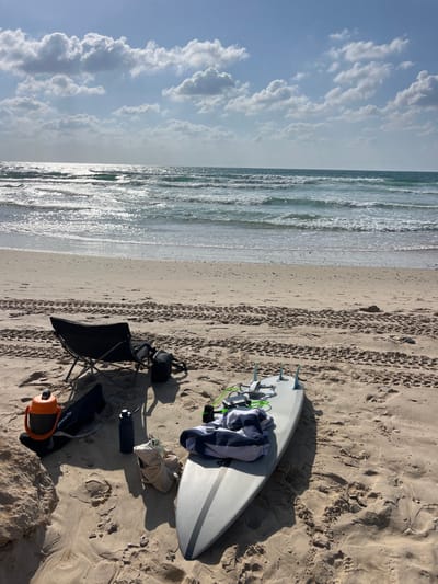 The image displays a beach scene at Beit Yanai, Israel, under a partly cloudy sky with bright sunlight. The foreground consists of light-colored sand marked with tire tracks and footprints. In the lower left, an orange insulated jug and a dark blue water bottle are visible, alongside a dark item of clothing or gear. A black foldable beach chair is positioned centrally, with a black bag near its base. To the right, a light-grey surfboard with a dark stripe and three fins rests on the sand. A white drone with four propellers is placed on the surfboard, which is also covered by a blue and white striped towel and has a green leash attached. The midground shows the Mediterranean Sea with moderate waves, reflecting sunlight across its surface. The background features a clear horizon line separating the sea from a blue sky dotted with numerous white cumulus clouds. No individuals are present in the scene.
