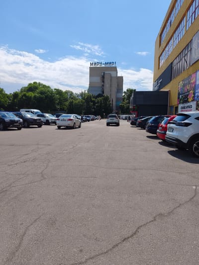 This is a daytime, eye-level, outdoor, full shot of a parking lot in front of a modern, yellow-and-gray building. The sky is blue with scattered white clouds. Tall green trees are visible behind the parking lot, and a tall, gray building with blue lettering on top is in the background. The parking lot is paved with asphalt and has visible cracks. Several cars of various colors and models are parked in the lot. The building on the right has a yellow facade with rows of windows and displays colorful advertisements. Text visible on the building includes "МИРУ-МИР!" which is also on the gray building, and advertisements for "СВІЖЕ М'ЯСО" (fresh meat) and "КАВА & ПУНДИКИ" (coffee & donuts). There are a few people visible walking in the parking lot. The overall impression is a sunny day at a commercial area.