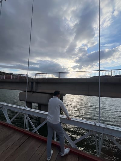 A man wearing gray trousers, a white top, and white shoes stands on the side of a bridge. He is holding onto the bridge's white metal railing. The bridge is over water. In the background, there is another bridge and a cloudy sky. The railing has a red stripe on the right side. There is a wooden surface on the right side. The location appears to be Tabaroucht, Morocco.