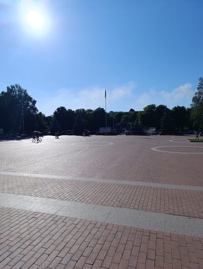 A wide, sun-drenched plaza paved with reddish-brown brick is the dominant feature of this image. The plaza is laid out in a pattern with parallel bands of gray gravel and lighter brick. In the mid-ground, several people are present. One individual is riding a bicycle, their silhouette outlined against the bright sky. Other figures are walking or standing in small groups, appearing as distant silhouettes.
In the background, a line of lush green trees forms a dense border. Behind the trees, a bright blue sky dominates, with the sun prominently positioned in the upper left quadrant, casting a strong glare and lens flare. A few wisps of white clouds are scattered across the sky. A flag pole with a flag is visible on the left side of the background. There are also several vehicles parked in the background, partially obscured by trees.
The scene appears to be set during the daytime, with clear, sunny weather. The bright sunlight suggests it could be a warm day. The overall mood is serene and open, with the vast plaza and clear sky conveying a sense of spaciousness. While no discernible text is clearly legible in the image, there are some signs or posters visible in the background among the trees. The presence of people engaged in leisurely activities like cycling and walking suggests a public space being utilized for recreation.