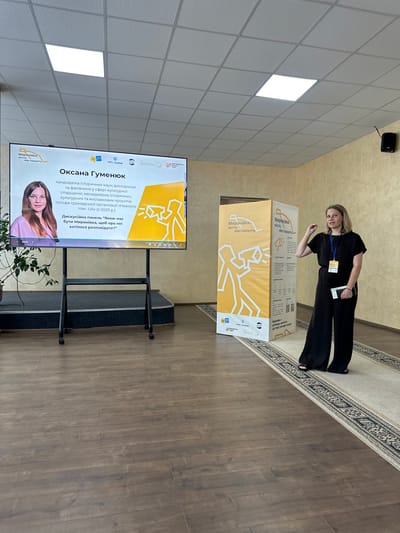 A woman stands to the right of a large, yellow display board, which features Ukrainian text and graphic designs. The woman is wearing a black outfit and a lanyard with an ID card. She is holding a dark object in her left hand. To her left, a large screen displays a presentation about "Oksana Gumeniuk," a candidate of historical sciences, researcher, and manager. The screen also shows the title of a discussion panel: "Myronivka - a city that speaks." In the background, the room has beige walls and a tiled ceiling with fluorescent lighting. The floor is covered with dark wooden planks and a patterned rug. The overall scene suggests an indoor event or presentation related to the city of Myronivka, Ukraine.
