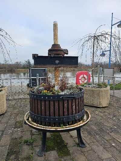 The scene is an outdoor public space in Bech-Kleinmacher, Luxembourg, under an overcast sky. In the foreground, a repurposed antique wine press functions as a planter. This object comprises a dark brown slatted wooden barrel, secured with black metal bands, resting on a three-legged black cast-iron base. A central, prominent light brown threaded screw extends vertically from the planter, topped by a heavy, dark horizontal metal beam. The barrel contains soil, sparse brown vegetation, small yellow flowering plants, and white specks consistent with frost or light snow. The ground is paved with grey rectangular pavers, interspersed with patches of green moss. In the mid-ground, a silver-colored metal railing runs along the edge of a wide, calm river. Beyond the railing, a red lifebuoy with white accents hangs visible. To the right, a tall blue lamppost with two luminaires and a slender leafless tree with weeping branches are present. A light-colored concrete octagonal planter is partially visible on the right side of the frame. The far background displays a distant line of leafless trees on the opposing riverbank. No individuals are visible in the scene.