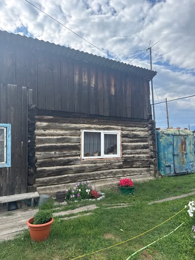 In Yabogan, Russia, a rustic, weathered wooden building with a corrugated metal roof stands under a cloudy sky. The building's lower section is constructed from horizontal logs, while the upper section is clad in vertical wooden planks. A white-framed window with sheer curtains is visible on the log section. To the left, a window with a light blue frame is partially visible. In front of the building, a wooden bench sits on a wooden deck. Flower pots containing pink and red flowers are arranged in the grassy area in front of the building, along with two buckets. To the right of the wooden building is a large, rusty, blue metal structure. Electrical wires are strung across the scene, supported by wooden poles in the distance. The ground is covered in green grass and a winding pathway made of wooden planks.