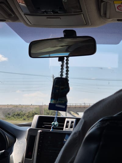 The image shows the interior of a car, looking forward through the windshield. The view outside the car reveals a bridge and a landscape with sparse vegetation under a clear sky with some clouds. Inside the car, a rearview mirror hangs from the windshield. Below the mirror, a car air freshener shaped like a tag with blue and white string and beads is suspended. The dashboard of the car is visible, featuring a touchscreen display, air vents, and buttons. A person's arm and shoulder are visible on the right side of the frame, suggesting the presence of a driver or passenger. The car appears to be on a road in or near Новоишим ауылдық округі, Kazakhstan, based on the provided information.