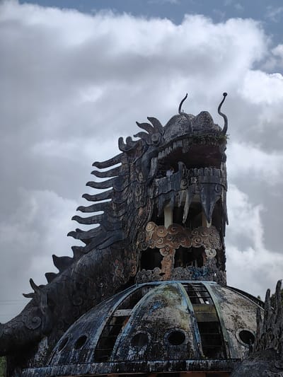 This image depicts a large, weathered dragon-shaped structure located in Kim Long Ward, Vietnam, likely part of an abandoned water park. The primary subject is a dark, dilapidated dragon head and upper body, rendered in concrete or similar material, dominating the upper two-thirds of the frame against an overcast sky with scattered white and gray clouds.
The dragon's head is shown with an open mouth, revealing jagged, tooth-like projections and an internal multi-tiered structure supported by columns. Within this internal structure, several indistinct human figures are visible on what appears to be a viewing platform or balcony. The dragon's head features two prominent, upward-curving antennae or horns and small patches of green vegetation, consistent with moss or ferns, growing on its surface. Its neck and back are detailed with textured, scale-like ridges.
Below the dragon's head and partially integrated with its lower structure is a large, dark, dome-shaped building. This dome exhibits significant signs of decay, with discolored surfaces, visible internal framework, and multiple circular openings or windows, some appearing broken or empty. The overall scene conveys a sense of abandonment and deterioration.
