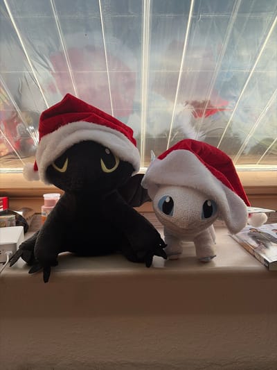 Two stuffed dragon toys, one black and one white, are positioned side-by-side on a ledge. Both toys are wearing red and white Santa hats. The black dragon is on the left, and the white dragon is on the right. Behind the toys, a window with a textured, translucent covering is visible. The ledge appears to be made of a light-colored material. To the left of the black dragon, there are various objects including a white electronic device and a small pink container. To the right of the white dragon, a magazine with a picture of people in winter clothing is lying flat. The lighting in the scene suggests it is daytime, with light filtering through the window. The image does not contain any information that would allow for the determination of its location in Göttingen, Germany.