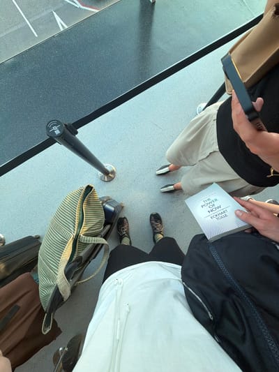 The image shows a group of people in a queue, likely at an airport or train station given the presence of luggage and stanchions. The perspective is from above, looking down at the individuals and their belongings. One person is holding a book titled "THE POWER OF NOW" by Eckhart Tolle. Another person is wearing light-colored shorts and black and white shoes, while another is in black pants and dark shoes. There are several pieces of luggage visible, including a striped duffel bag and a black backpack. The floor is a light gray, speckled surface, and a dark grey barrier or counter runs along the left side of the frame. The lighting suggests it is daytime, possibly indoors with artificial lighting or near a window. The overall mood is neutral, with people waiting. There is some visible text on the book and on a circular tag attached to one of the stanchions, but it is largely illegible.