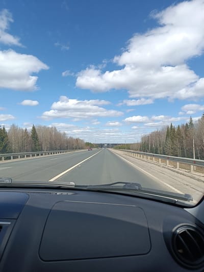 The image depicts a view from inside a car, looking out onto a highway. The dashboard of the car is visible in the foreground. Beyond the windshield is a straight stretch of road with multiple lanes. A dividing line separates the lanes. Guardrails run along the edges of the highway, separating it from forested areas on either side. The trees appear to be a mix of deciduous and evergreen varieties, with many deciduous trees still bare, suggesting it is early spring. In the distance, other vehicles can be seen on the road. The sky is blue with scattered white clouds. The scene suggests travel or commuting on a rural highway. There is no evidence to specifically indicate the location is in the city of Ленинск, Russia.