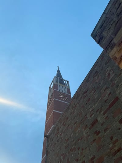The image presents a dramatic, upward-looking perspective of a tall clock tower, partially framed by an imposing overhead structure, against a clear blue sky.

The central subject is a clock tower, constructed from reddish-brown brick or similar material, adorned with lighter-colored structural accents around its corners and at the level of the clock faces. The tower features a dark, pointed spire capped with a small cross, indicating it likely belongs to a church or a significant public building. At least one large, round clock face is clearly visible on the tower's side, marked with radial lines to denote hours, though no clock hands are distinctly visible to tell the time. Another clock face is partially visible further down. Square or rectangular openings are also incorporated into the tower's design near its top.

The view of the tower and sky is framed by a substantial, dark, and textured architectural element that stretches across the top of the image. This element appears to be the underside of a large overhang, archway, or bridge, constructed from rough-hewn stone or brickwork, adding depth and a sense of enclosure to the composition.

The background is a vast expanse of bright, clear blue sky, suggesting good weather during the day. A subtle, elongated band of lighter blue or very thin cloud can be discerned towards the lower right portion of the sky, adding minor variation to the otherwise uniform azure. The lighting indicates it is daytime, possibly morning or afternoon.

The photograph captures an interesting interplay between modern and historical architectural elements in what is contextualized as Pforzheim, Germany, emphasizing the scale and design of the structures. No explicit text is visible within the frame, apart from the markings on the clock face.
