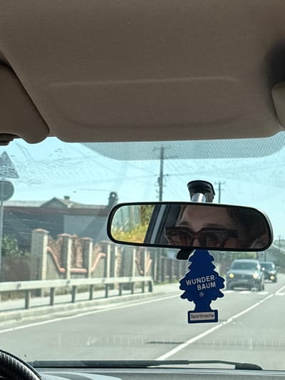 The image, taken from the interior of a car, shows a view through the windshield. The windshield has streaks and smudges. A rear-view mirror is visible, reflecting the upper half of a person wearing sunglasses. Below the mirror, a blue tree-shaped car air freshener labeled "WUNDER-BAUM" and "Sportfrische" hangs. Outside the car, a road with lane markings is visible, with a white guardrail and a decorative fence with brick pillars. Buildings are visible in the background. Two cars are driving on the road ahead. The environment appears to be urban or suburban. The image location is identified as Drohovyzh, Ukraine.