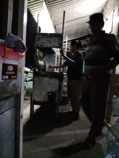 The image depicts an outdoor or semi-outdoor cooking scene at night in Juwana, Indonesia. In the mid-ground, a female individual with dark hair pulled back is actively cooking. She wears a dark shirt and light-colored pants, and is seen stirring a large metal wok or pan placed over a visible flame on a makeshift stove structure. This structure consists of a metal frame and what appears to be a concrete or plaster-like base.
In the immediate foreground, to the right, a male individual in dark clothing, including a cap, is partially visible and out of focus, appearing to walk or stand in profile. To the left foreground, a white plastic storage container with a pink lid is present, topped with a crumpled grey cloth. A "SHINPO HANDY CD 15" sticker is affixed to the container. Part of a metal post or structure stands next to it. The ground is paved. Overhead, a corrugated metal roof is visible, suggesting a covered area. The scene is dimly lit by artificial light sources, casting shadows and contributing to the overall dark environment. Transparent plastic bottles are partially visible on the far right of the foreground.