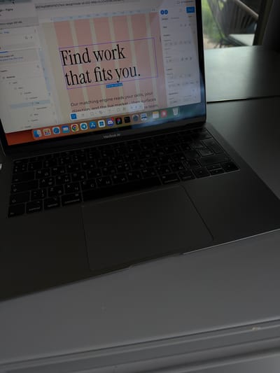 The image shows a MacBook Air laptop open, displaying a design project in Figma. The design features large text reading "Find work that fits you." in a bold font, with pink columns behind the text. Below this is the text: "Our matching engine reads your skills, your direction, and the live market - then surfaces he team." The screen also shows the Figma design interface with various tools and panels. The keyboard of the MacBook Air is visible, with Cyrillic characters on the keys. The laptop is placed on a surface near a window with a view of greenery outside.