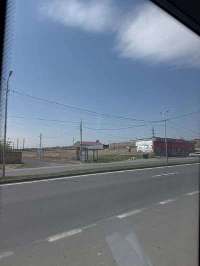This image, taken from inside a vehicle, captures a roadside scene in Ptghunk, Armenia, under a clear, bright blue sky with a large, diffuse white cloud. The image is oriented vertically (originally landscape, rotated 90 degrees clockwise).
The right side of the frame features a paved road with distinct white dashed lane markings. A narrow grass verge separates the road from the adjacent land. Numerous utility poles with a complex network of overhead power lines crisscross the middle and left portions of the image.
In the midground, a low brick wall runs parallel to the road. Behind it, a small building with a red roof and what appears to be a bus stop or shelter is visible. Further along, a large, prominent red building stands out with multiple windows and extensive signage. Visible text on this building includes "ԳՐԱՆԴ ՔԱՐԵՐ" (Grand Karer), "MAX SHINE", "24/7", and "ԱՄԲՈՂՋԱԿԱՆ ԼՈՒԾՈՒՄՆԵՐ" (Complete Solutions). A white car is parked in front of this red building. Below it, a billboard displays "TOTAL" along with Armenian text reading "ԱՎՏՈԱՊԱՀՈՎԱԳՐՈՒԹՅՈՒՆ" (Auto Insurance) and "ՊԱՐՏԱԴԻՐ ԱՊԱՀՈՎԱԳՐՈՒԹՅՈՒՆ" (Mandatory Insurance).
The top-left corner (originally bottom-left) shows a reflection, likely from a vehicle's dashboard or window frame. The overall impression is that of a commercial or semi-urban area alongside a main road, photographed during daytime under sunny weather conditions.