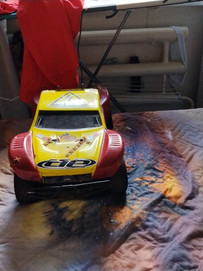 This media file shows a yellow and red remote-controlled car resting on a patterned blanket. The car is the primary subject, and it is positioned in the foreground, angled slightly towards the viewer's left. It has a prominent "d2" decal on the front and appears to be a toy model, possibly a buggy or off-road vehicle. Behind the car, a red piece of fabric is draped, possibly clothing or a curtain, and behind that, a metallic structure, possibly a drying rack or shelving unit, is visible. The background is dimly lit, suggesting an indoor setting. The blanket the car is on has a colorful, abstract pattern in shades of orange, blue, and purple, with visible stitching. There is no clear indication of time of day or weather. The setting appears to be a domestic environment. No people are visible in the frame.