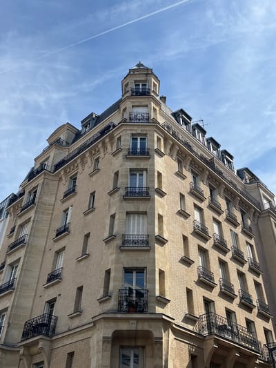 This is a low-angle, full-frame, eye-level exterior shot of a multi-story building with a light brown brick facade and stone accents. The building has multiple windows with black wrought-iron balconies. The windows are a mix of open and closed, with some featuring white shutters. The roofline is visible, with several dormer windows and a small turret with a pointed roof. The sky is blue with some wispy white clouds and a faint contrail. The building appears to be located in Le Kremlin-Bicêtre, France, based on the architectural style. There are no people visible in the image. The overall impression is that of a historic Parisian-style apartment building.