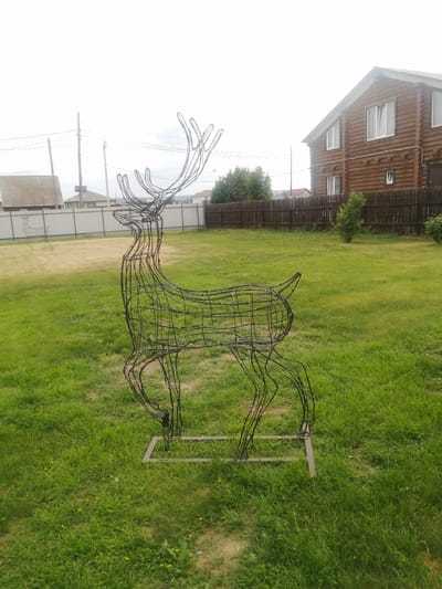 A wireframe deer sculpture stands on a green lawn in Yemelyanovo, Russia. The sculpture is a life-size silhouette of a deer, constructed from dark wire, with intricate detailing in its antlers and legs. Behind the deer, a weathered wooden fence borders a grassy yard, and further back, a two-story wooden house with white-trimmed windows is visible. The sky is overcast, suggesting a cloudy day.